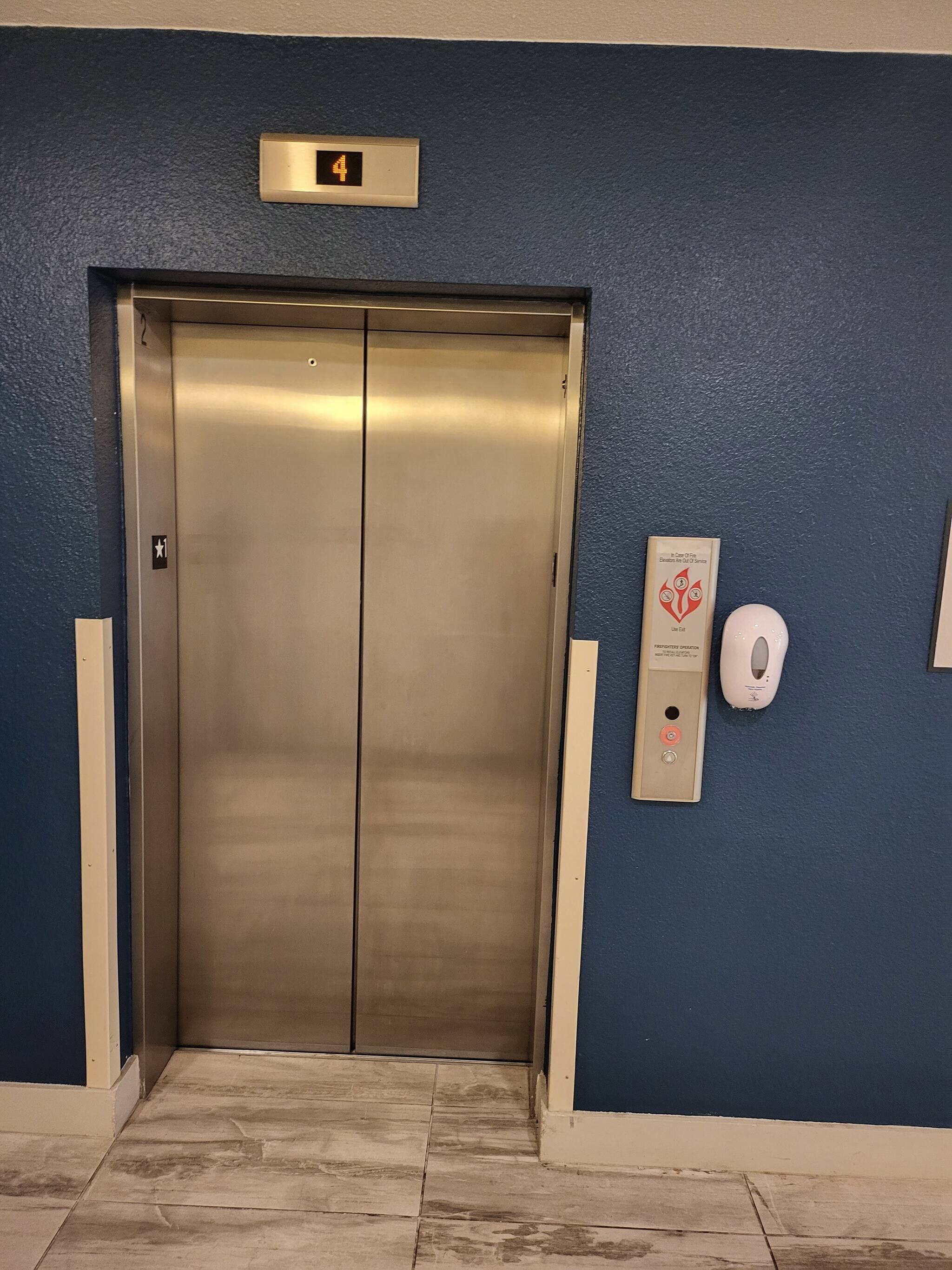 Un ascensor amb un pantalla digital que mostra el número 4, un dispensador de gel hidroalcohòlic i un senyal que indica 'Use Exit'.