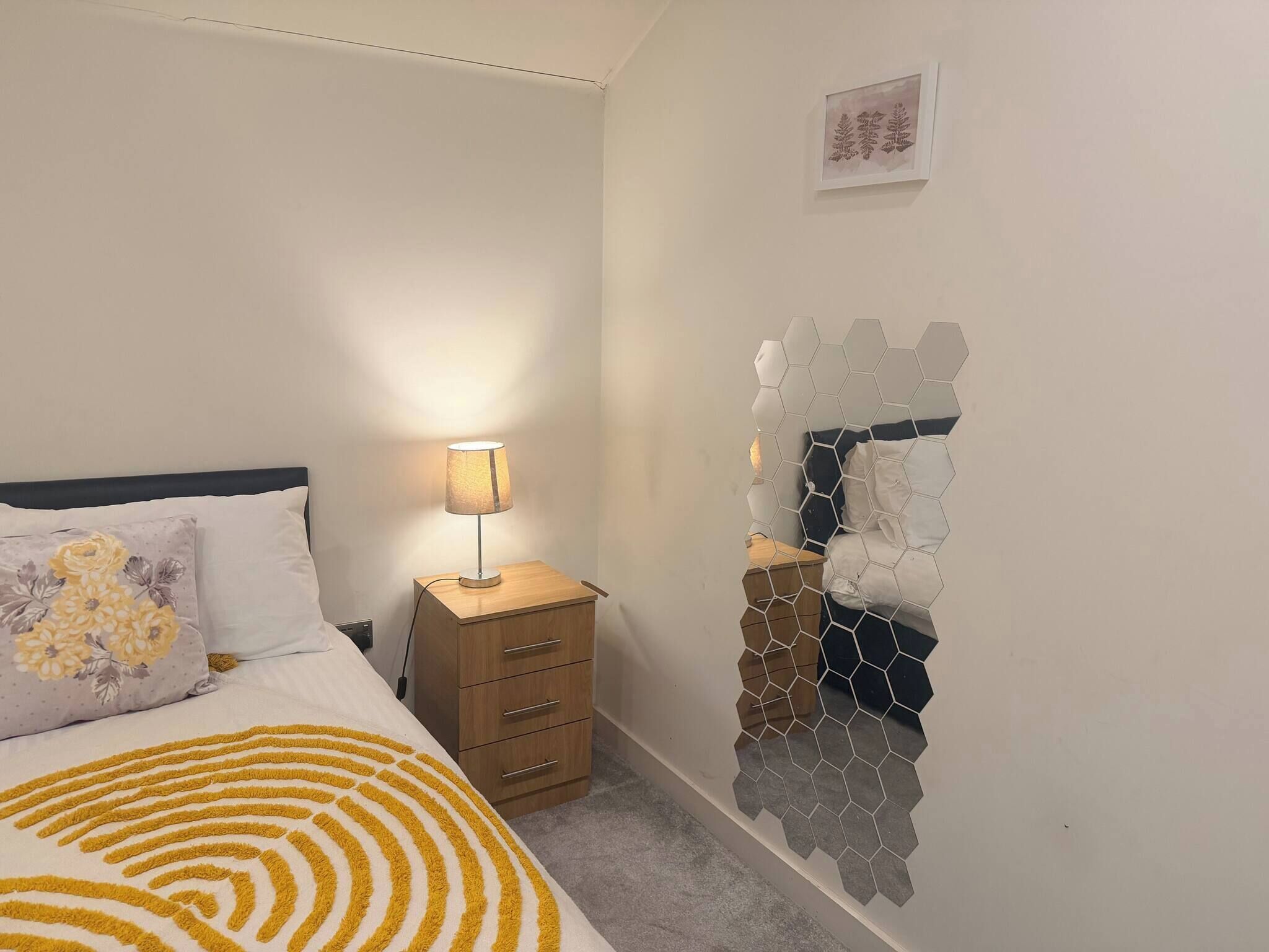 A bedroom with a bed, a bedside table with a lamp, a dresser, and a wall with hexagonal tiles.