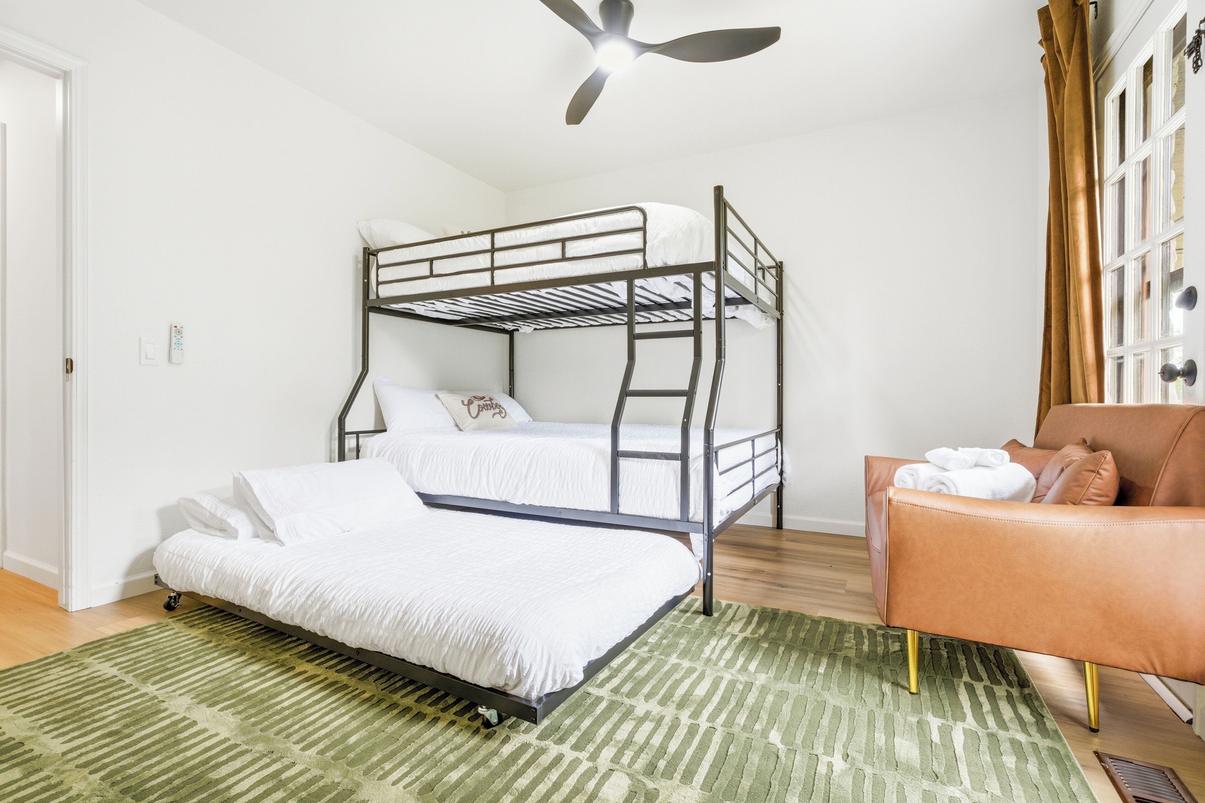 Un lit superposé, un tapis à rayures vertes, un fauteuil en cuir marron et un ventilateur de plafond.
