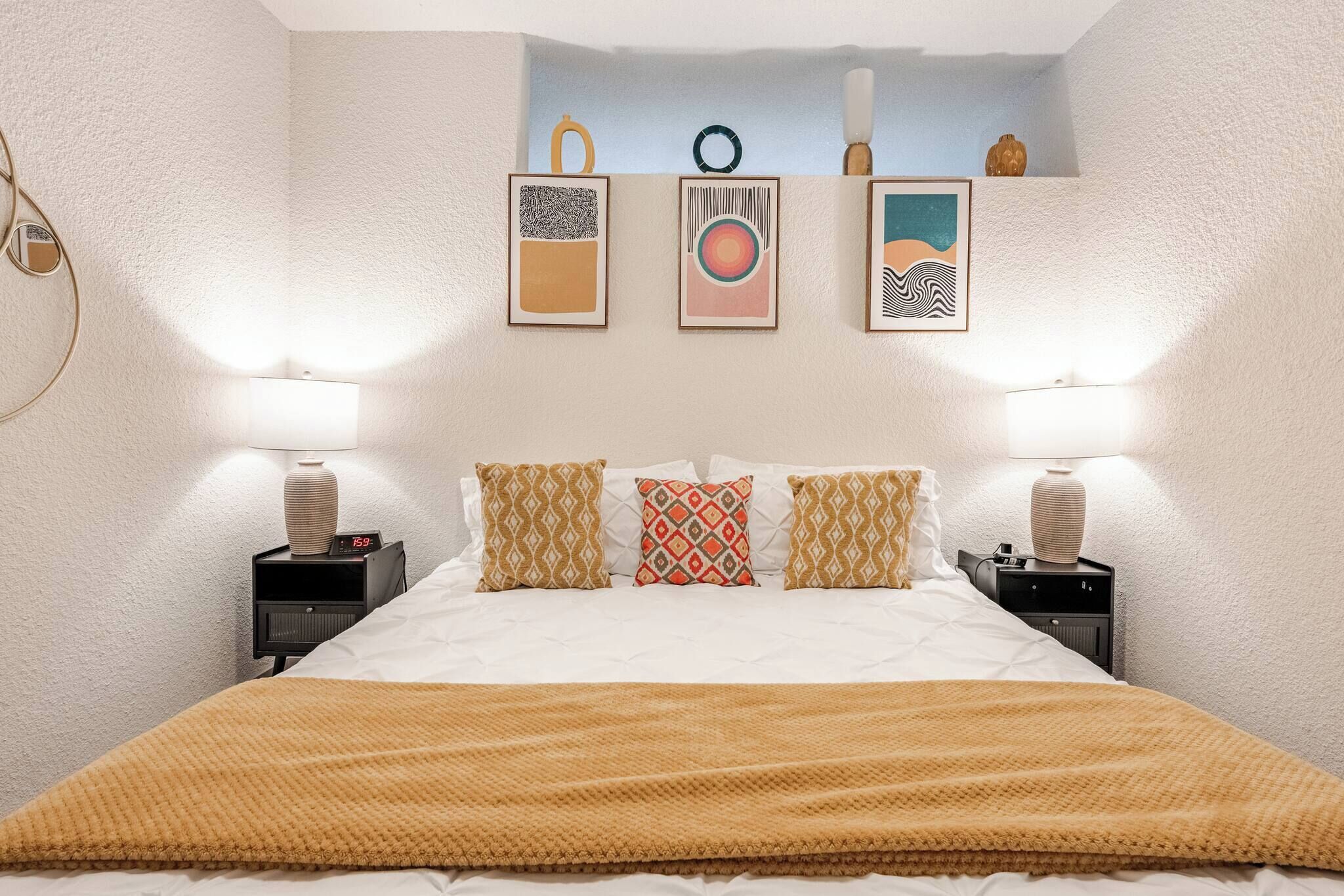 Une chambre à coucher bien rangée, avec un lit, des coussins à motifs, des tables de chevet avec des lampes et des œuvres d’art accrochées au mur.