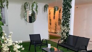 Une pièce avec un tapis vert synthétique, des chaises noires, une petite table sur laquelle est posée une plante, et des plantes décoratives suspendues.
