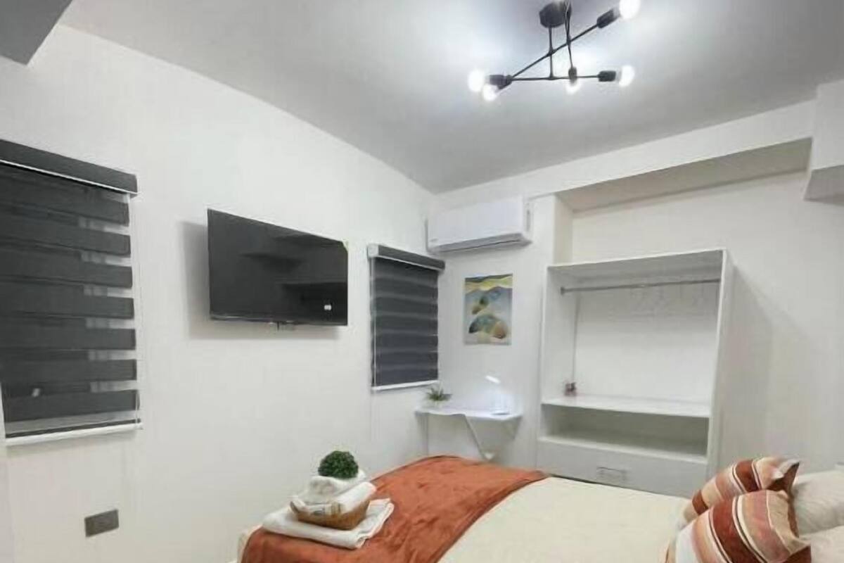 A modern bedroom with a bed, a flat-screen TV, a ceiling light fixture, and a window with blinds.