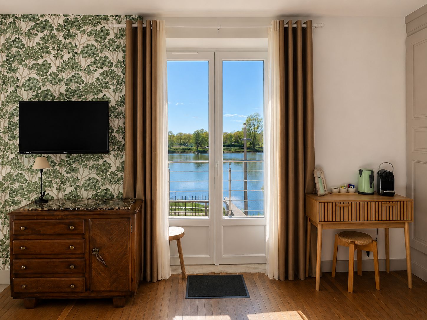 A room with a wooden dresser, a television, a desk with a chair, and a view of a lake through a glass door.