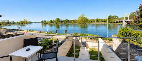 Een terras met een tafel en stoelen met uitzicht op een rustige rivier, met een brug en groen in de verte.