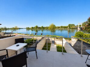Una terraza con mesa y sillas con vista a un río tranquilo, con un puente y vegetación al fondo.