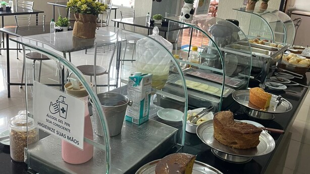 A buffet area with various food items, a sign indicating alcohol gel availability, and a glass display case. - Hotel Pousada das Palmeiras (Colider)