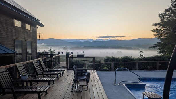 Eine Holzterrasse mit Sitzgelegenheiten im Freien, einem Pool und Blick auf nebelverhangene Berge.