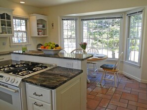A kitchen with a gas stove, a bowl of fruit, a dining table with chairs, and large windows. - Room for your kids, your parents or your sister or brother and their family. (Yarmouth)