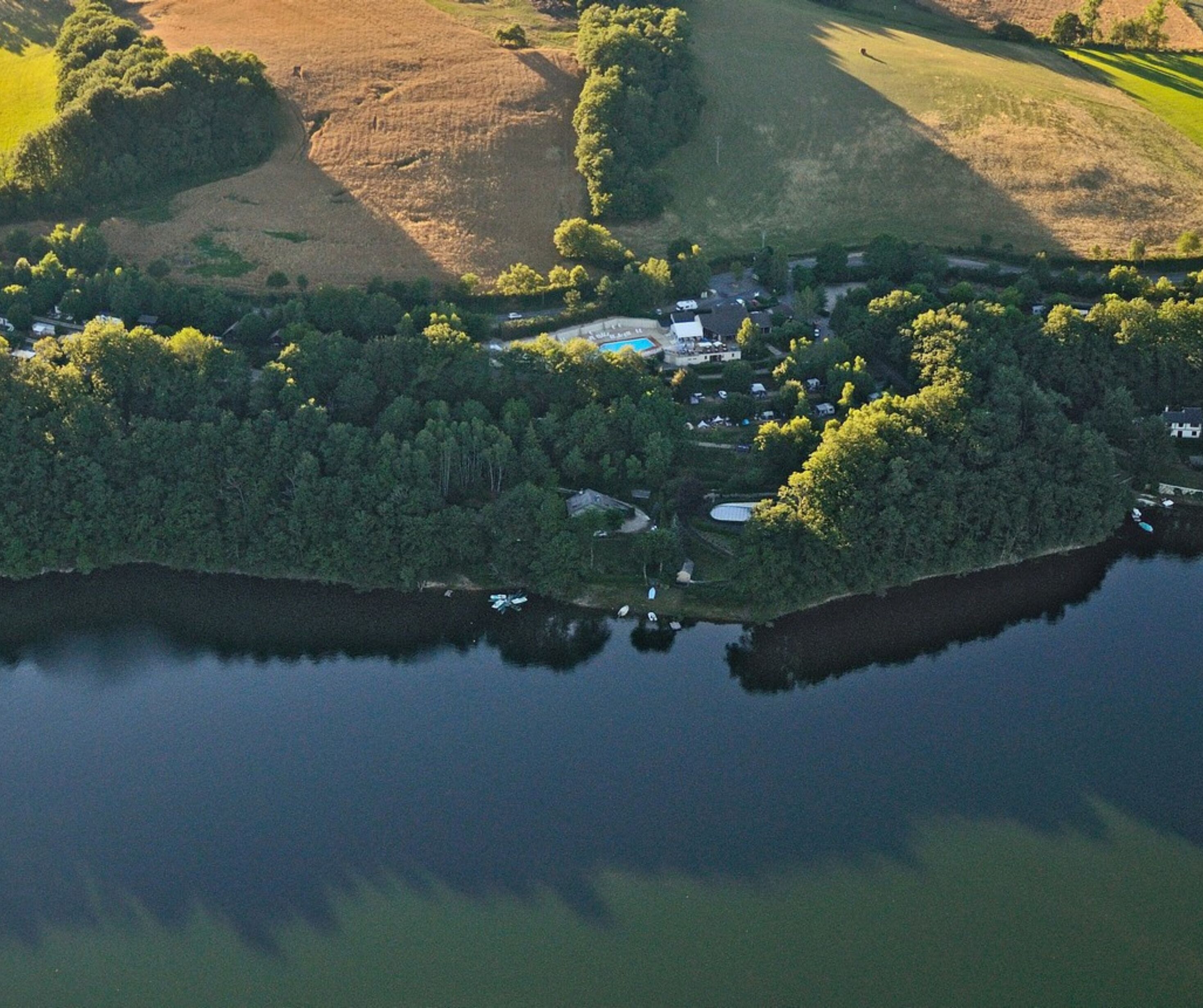 An aerial view of a lakeside property with a swimming pool, surrounded by dense trees and a small cluster of buildings.