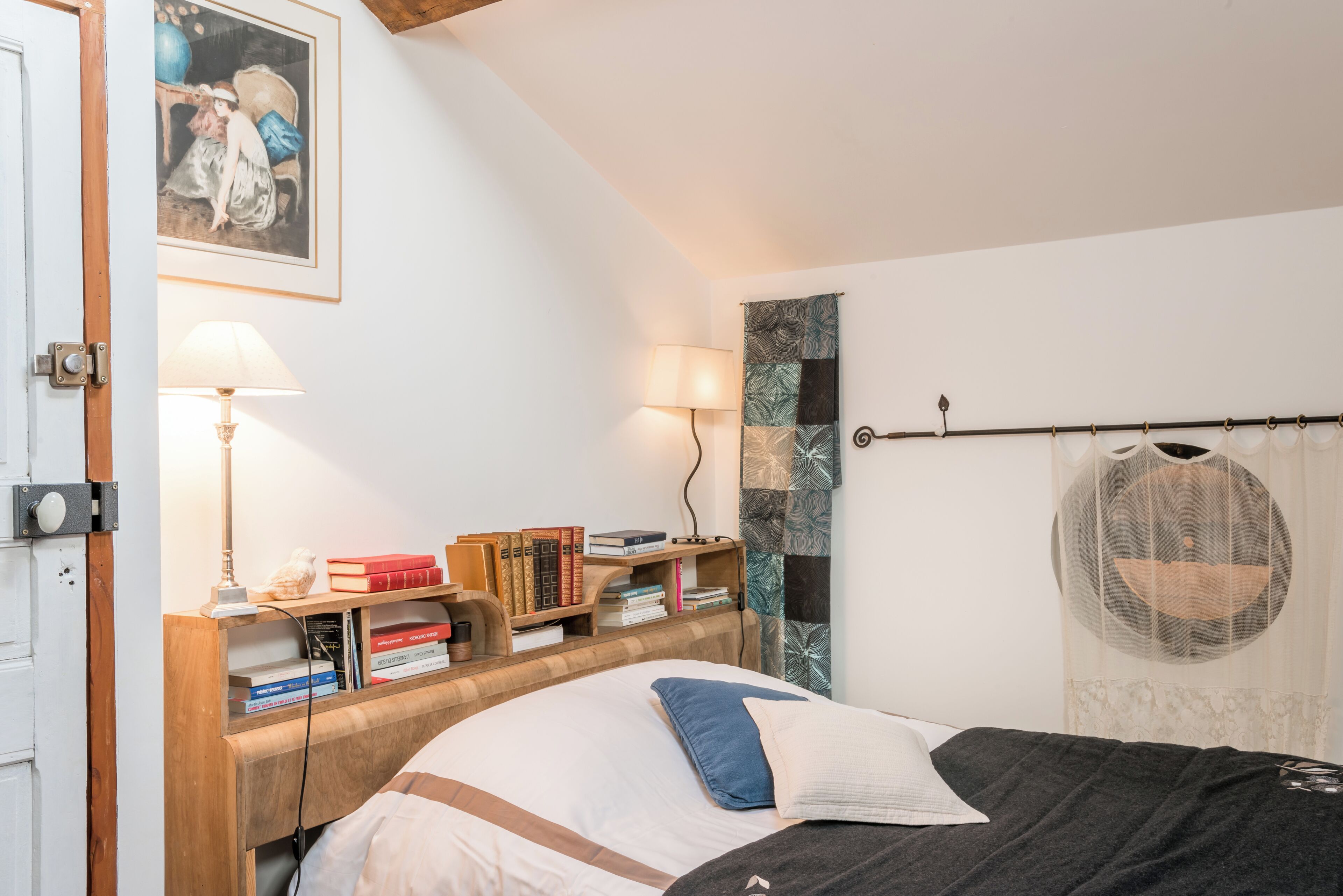 A bedroom with a wooden headboard, a bed with pillows, a bedside table with books, a lamp, and a wall with a framed picture.