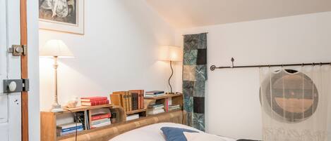 A bedroom with a wooden headboard, a bed with pillows, a bedside table with books, a lamp, and a wall with a framed picture.
