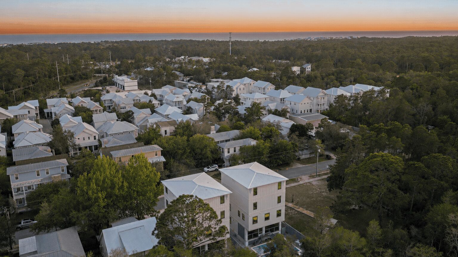 A residential area with multiple houses, some with visible balconies, surrounded by trees and a view of the ocean at sunset.