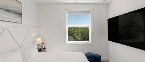 A bedroom with a bed, a nightstand, a lamp, a window with a view, and a TV.