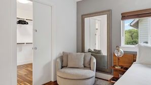 A bedroom with a bed, a round armchair, a large mirror, a bedside table with a lamp, and a view of greenery outside. - Govalle Glow by Avantstay East Austin Retreat Firepit Near Downtown + Nightlife (Austin)