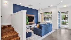A modern living room with a blue accent wall, a flat-screen TV, a blue sofa, and a staircase. - Govalle Glow by Avantstay East Austin Retreat Firepit Near Downtown + Nightlife (Austin)