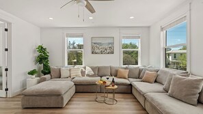 A modern living room with a sectional sofa, a ceiling fan, large windows, and a view of the outdoors. - Grayton Dreams by Avantstay Sleeps 32 Group Retreat Pool + Game Room + Golf Carts (Santa Rosa Beach)