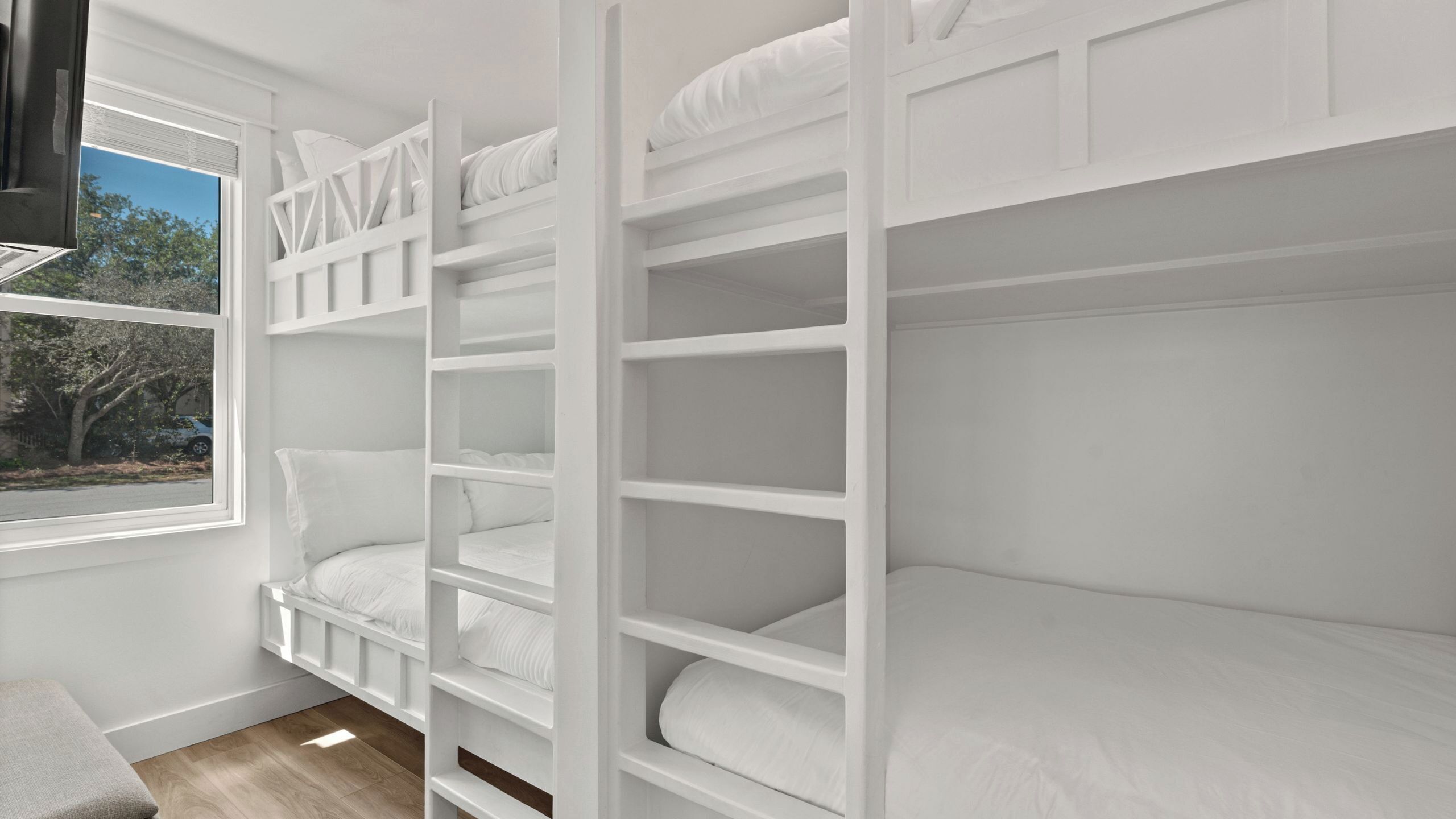 A bunk bed with white bedding, a window with a view of trees, and a wooden floor.