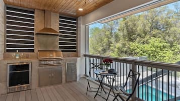 Une cuisine extérieure équipée d’un barbecue, d’un four et d’un porte-verres à vin, avec vue sur une piscine et des arbres.