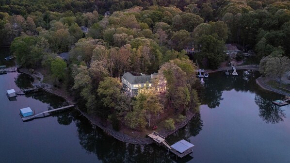 An aerial view of a lakeside property with a house surrounded by dense forest, docked boats, and a small dock with a covered area. - Montclair Manor by Avantstay Lakefront Retreat Dock + Hot Tub + Firepit + Game Room (Mooresville)