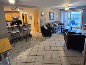 A living room with a sofa, chairs, a dining area with a table and chairs, a kitchen with a microwave, and a large window with a view. - Lightkeeper's Nest ~ Condo + Pool Hot Tub & Sauna (Port Clinton)
