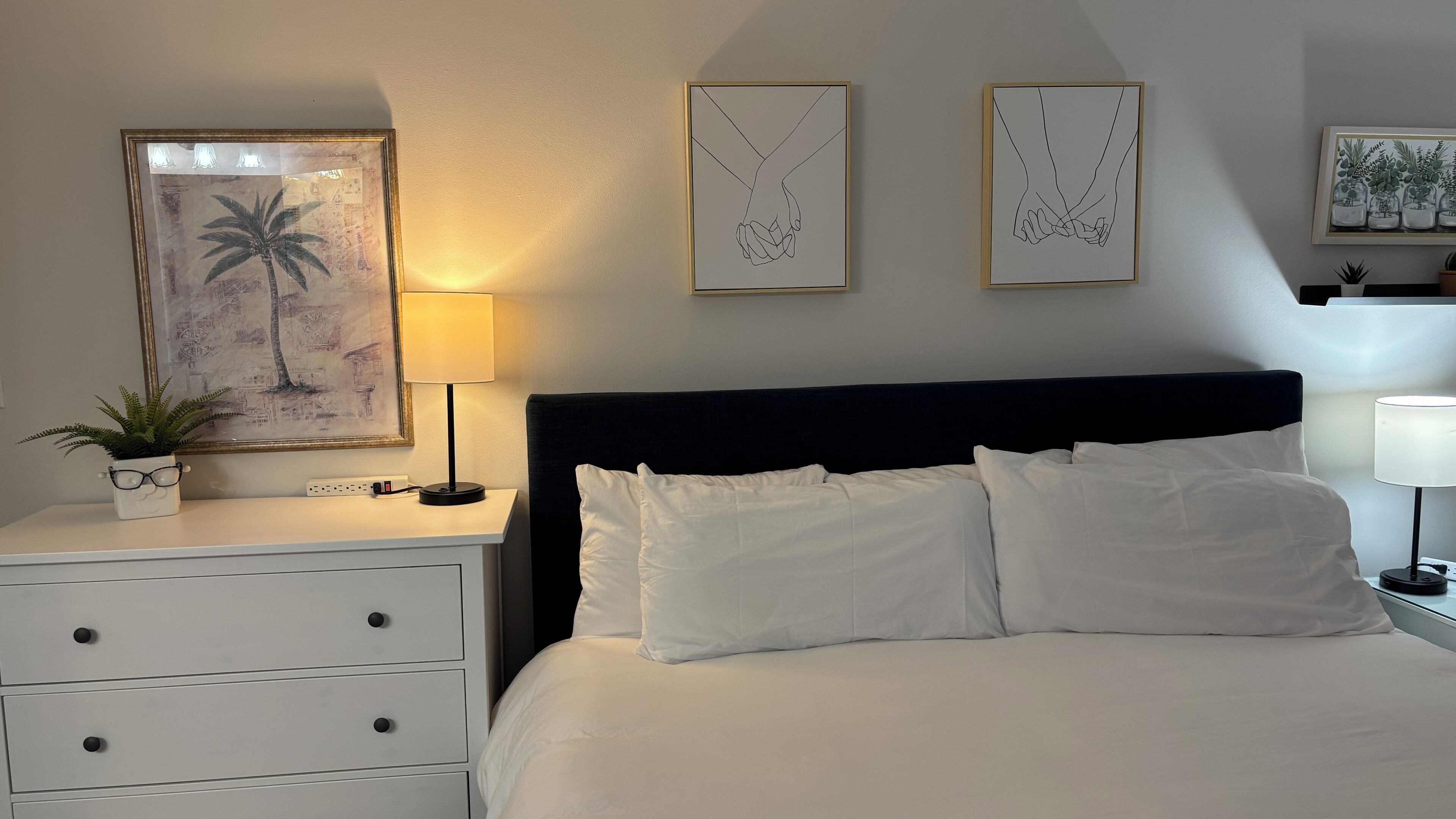 Hotellihuone, jossa on sänky, yöpöydät, lamppu, kehystetty kuva käsistä ja huonekasvi.