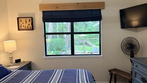 Ett sovrum med en säng, ett sängbord med en lampa, en byrå, en tv som är monterad på väggen och ett fönster med utsikt över en trädgård.