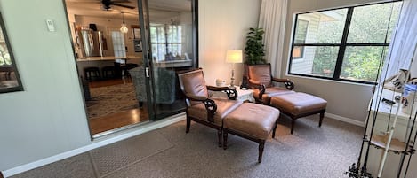Een kamer met een schuifdeur naar een eethoek, twee leren fauteuils, een kleine tafel met een lamp, en een groot raam met uitzicht.