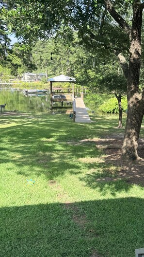 A grassy area with a wooden dock leading to a gazebo by a calm lake, surrounded by trees.
