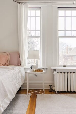 A bedroom with a bed, a small table with a lamp, a radiator, and a window with curtains. - Studio at Humboldt House (Denver)