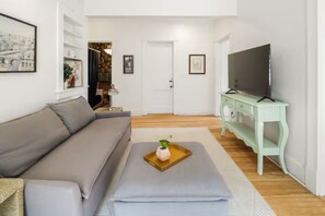 A living room with a sofa, a television on a stand, a door, and a window with curtains. - Park View Apartment at Humboldt House (Denver)