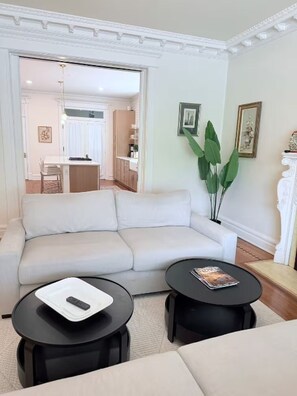 Ein Wohnzimmer mit einem weißen Sofa, zwei schwarzen Couchtischen, einer Fernbedienung und einer Zeitschrift auf einem Tisch. Eine Tür führt zu einer Küche und einem Essbereich.