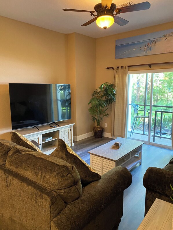 A living room with a flat-screen TV, a ceiling fan, a potted plant, and a view of the outdoors through a large window. - Bahama Bay Resort, 3 bedroom lake/pool view with 75 inch TV. Ground floor. (Davenport)