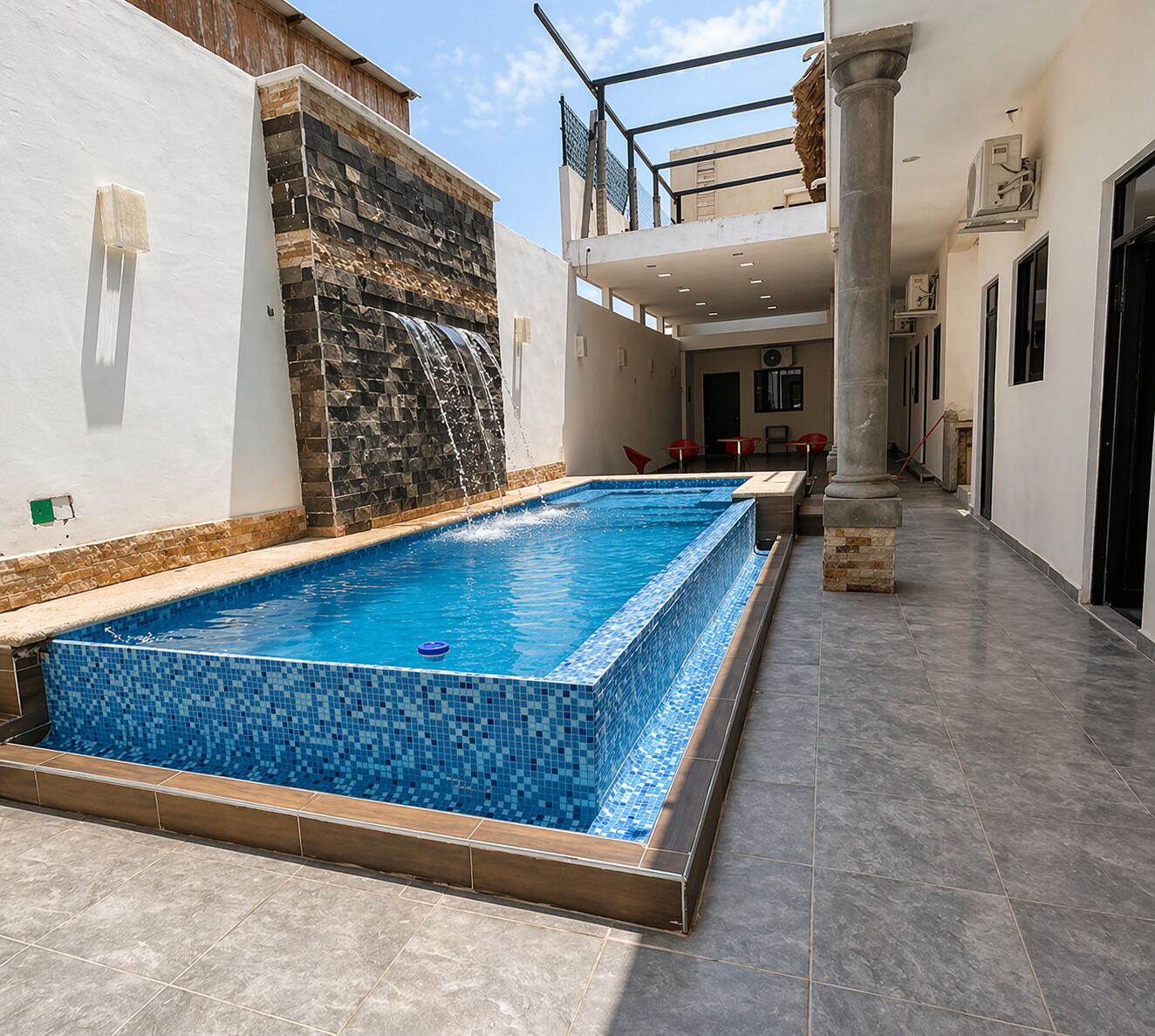 Ein rechteckiger Swimmingpool mit blauen Mosaikfliesen, einer Steinwand und einem gefliesten Boden.