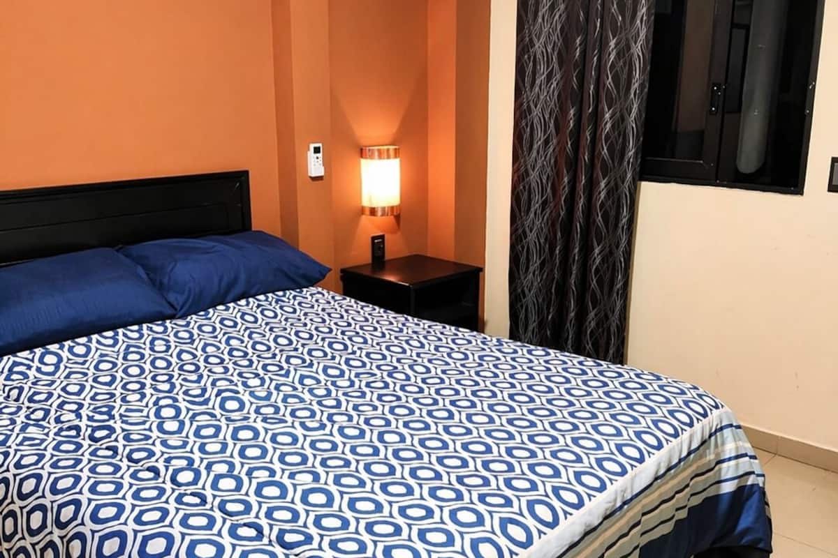 Une chambre d’hôtel comprenant un lit, une table de chevet, une lampe, un appareil de climatisation et une fenêtre avec des rideaux.
