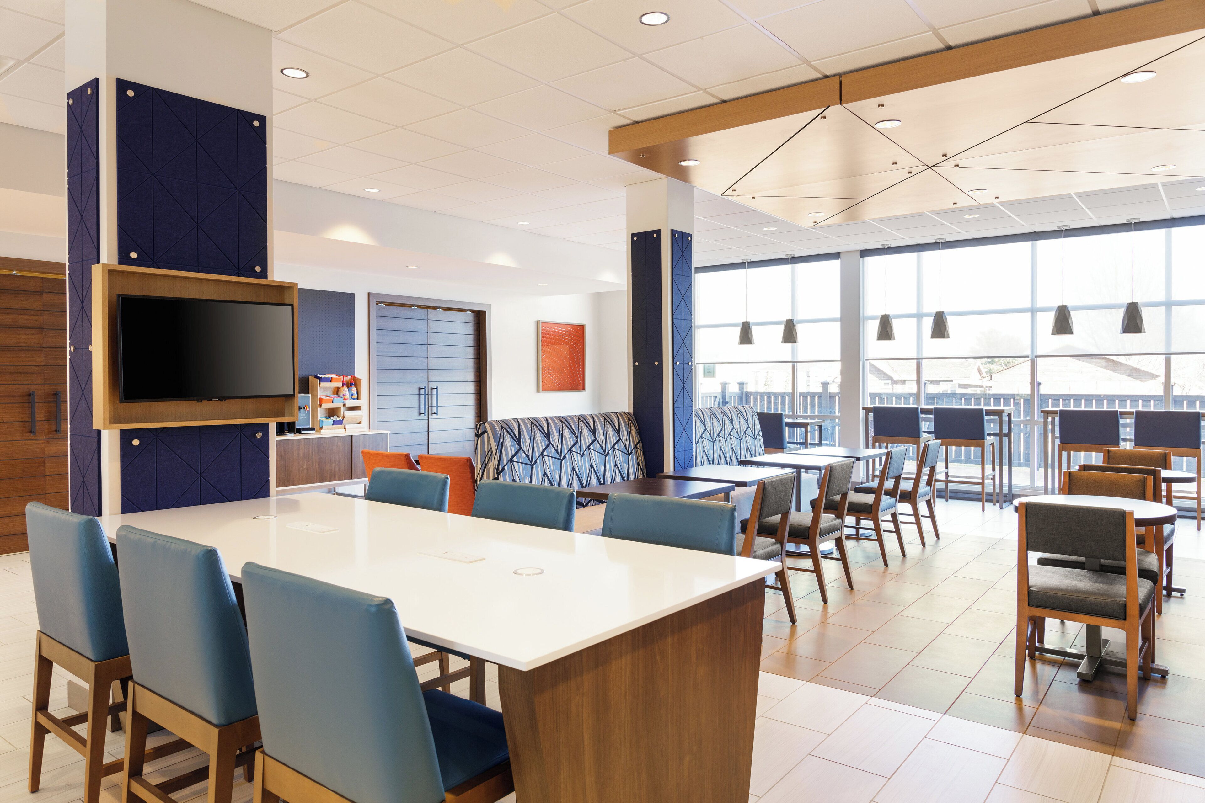 A modern hotel lobby with a large dining area, a flat-screen TV, and large windows.