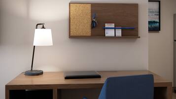 A modern office desk with a laptop, a desk lamp, a corkboard with keys and a sign, and a framed picture on the wall.
