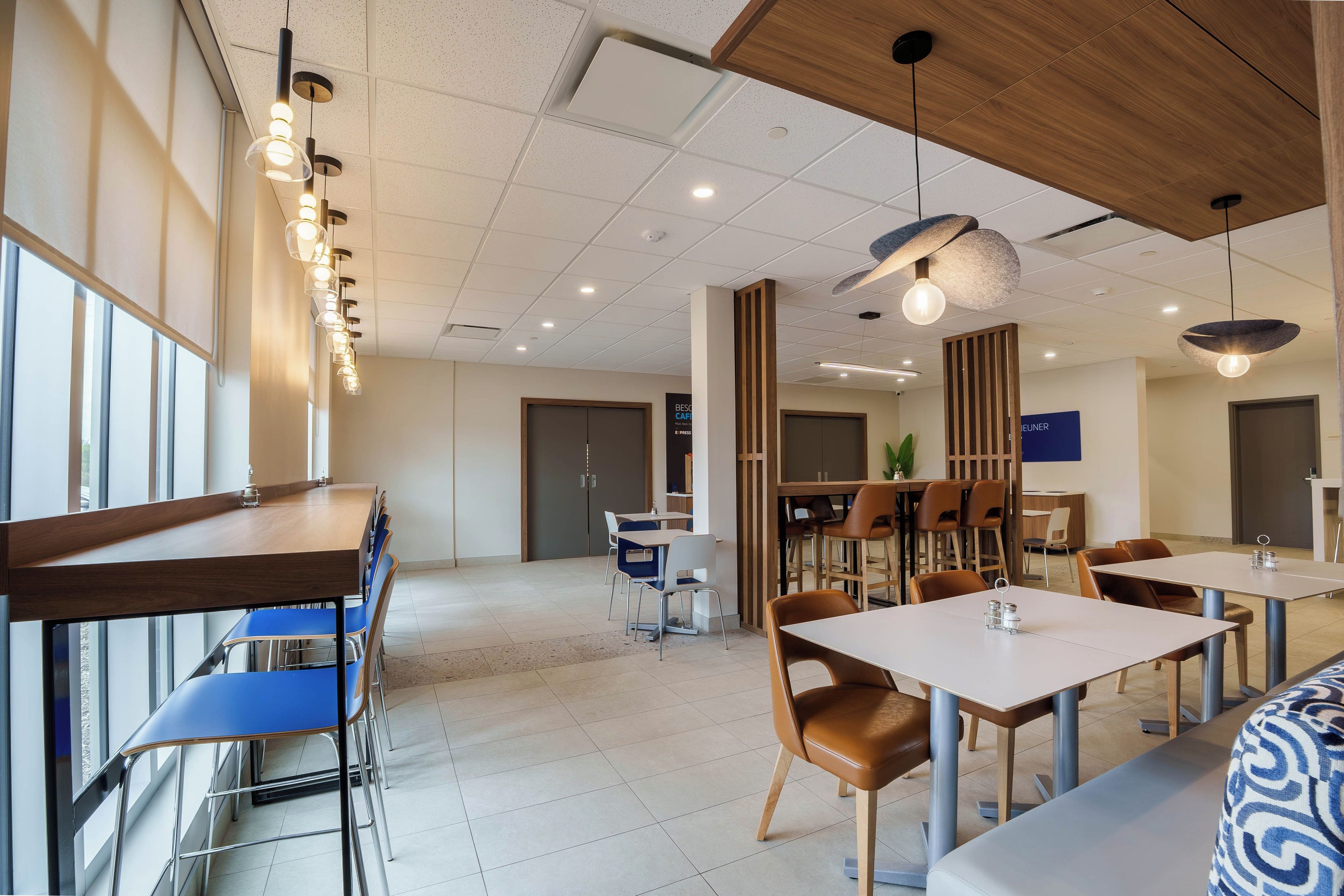 Un espace de restauration moderne, doté de plusieurs tables et chaises, d’un plafond pourvu d’un éclairage encastré et offrant une vue sur l’extérieur grâce à de grandes fenêtres.