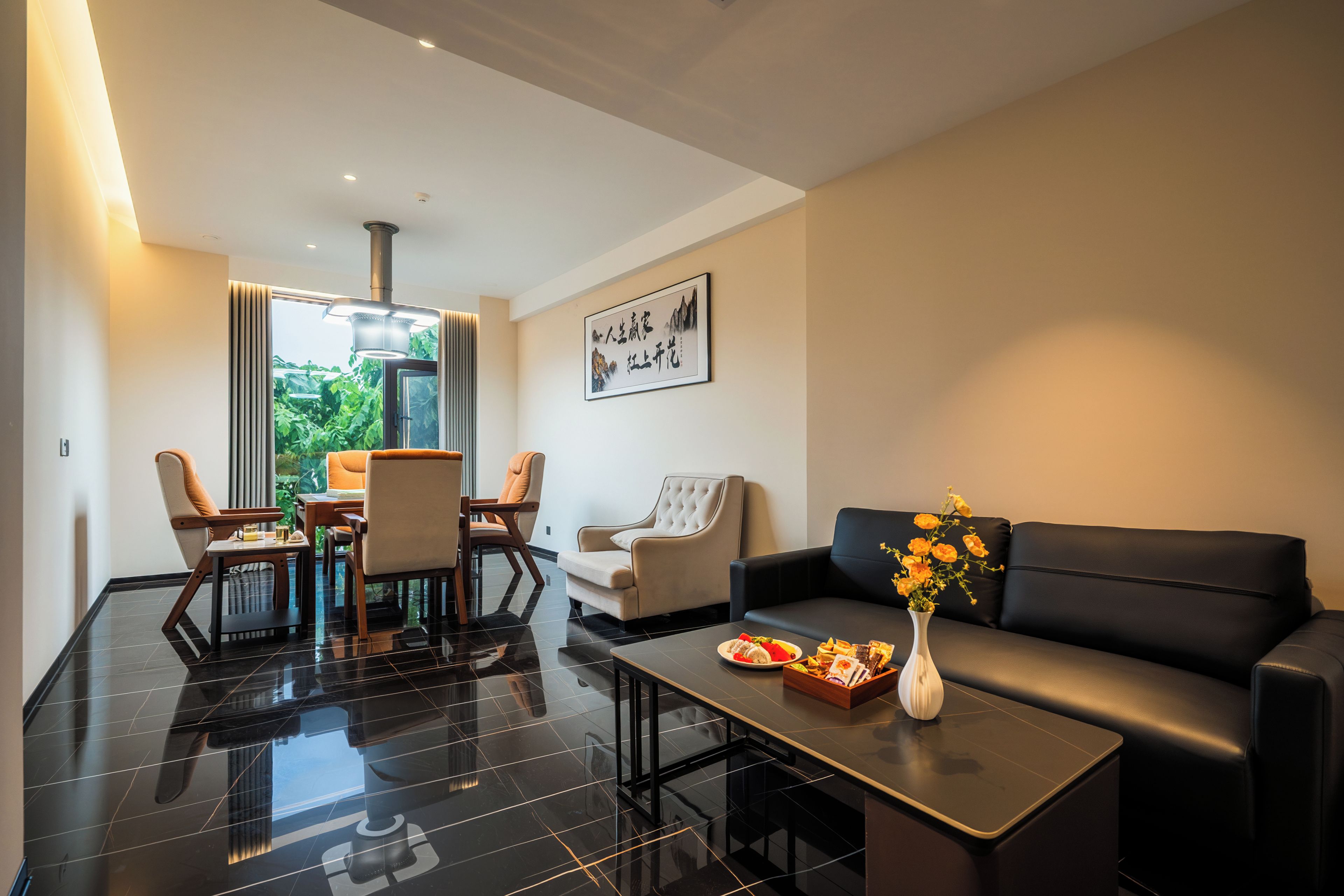 A modern living room with a black leather sofa, a dining area with wooden furniture, and a large window offering a view of greenery.