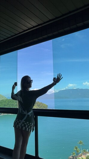 A person standing by a large window with a view of a body of water and mountains. - 3-Bedroom Apartment in Porto Real | Ocean View + Full Resort Amenities (up to 10 people) (Mangaratiba)