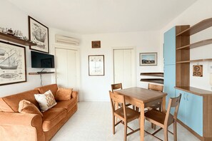 Una habitación con una mesa de comedor de madera, sillas, un sofá con cojines, un televisor, cuadros en las paredes y un mueble azul.