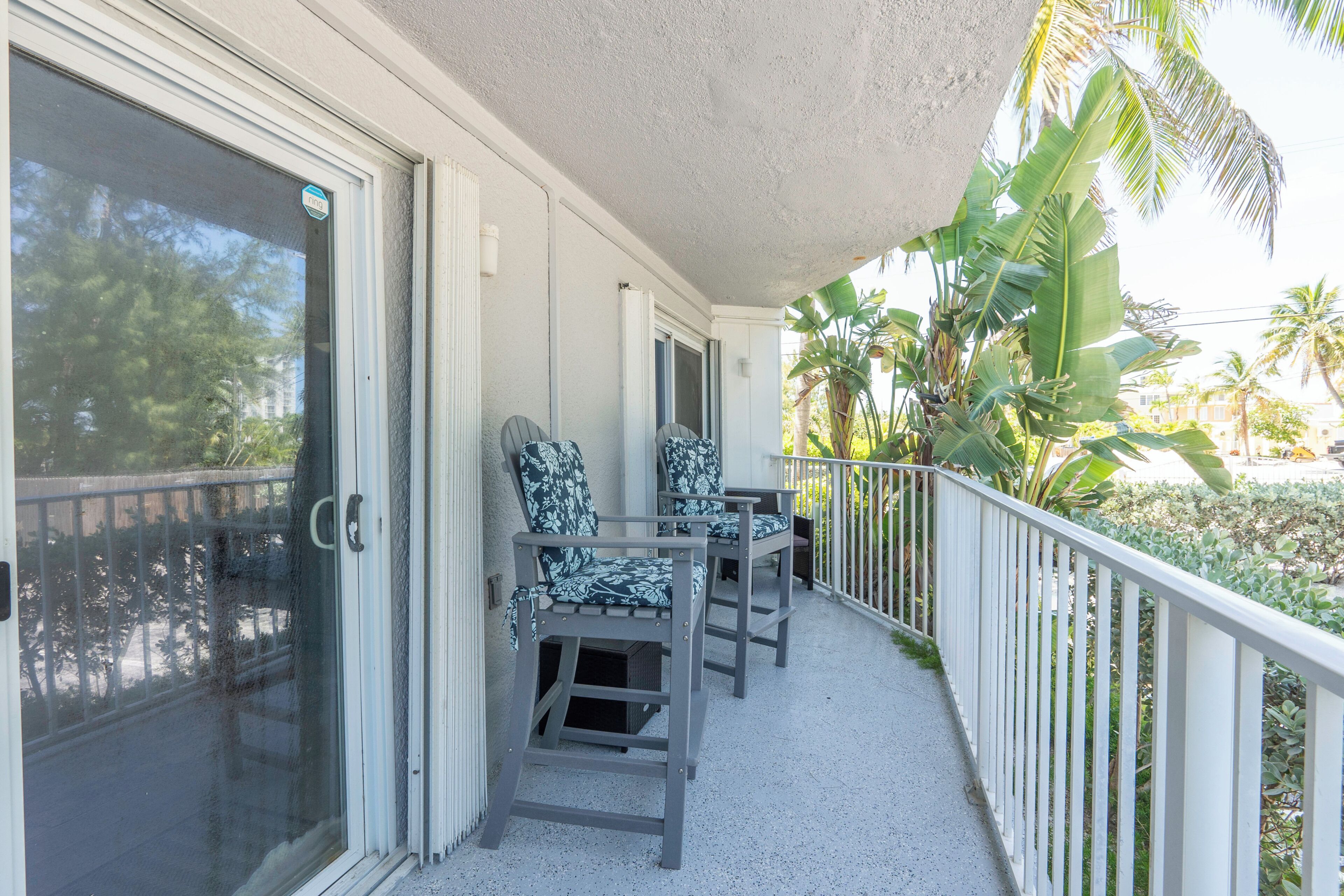A covered balcony with two chairs, a white railing, and a view of palm trees and greenery.