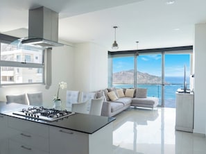 Eine moderne Küche mit Gasherd, eine Essecke mit Stühlen, eine Sitzecke mit Sofa und große Fenster mit Blick auf das Meer und die Berge.