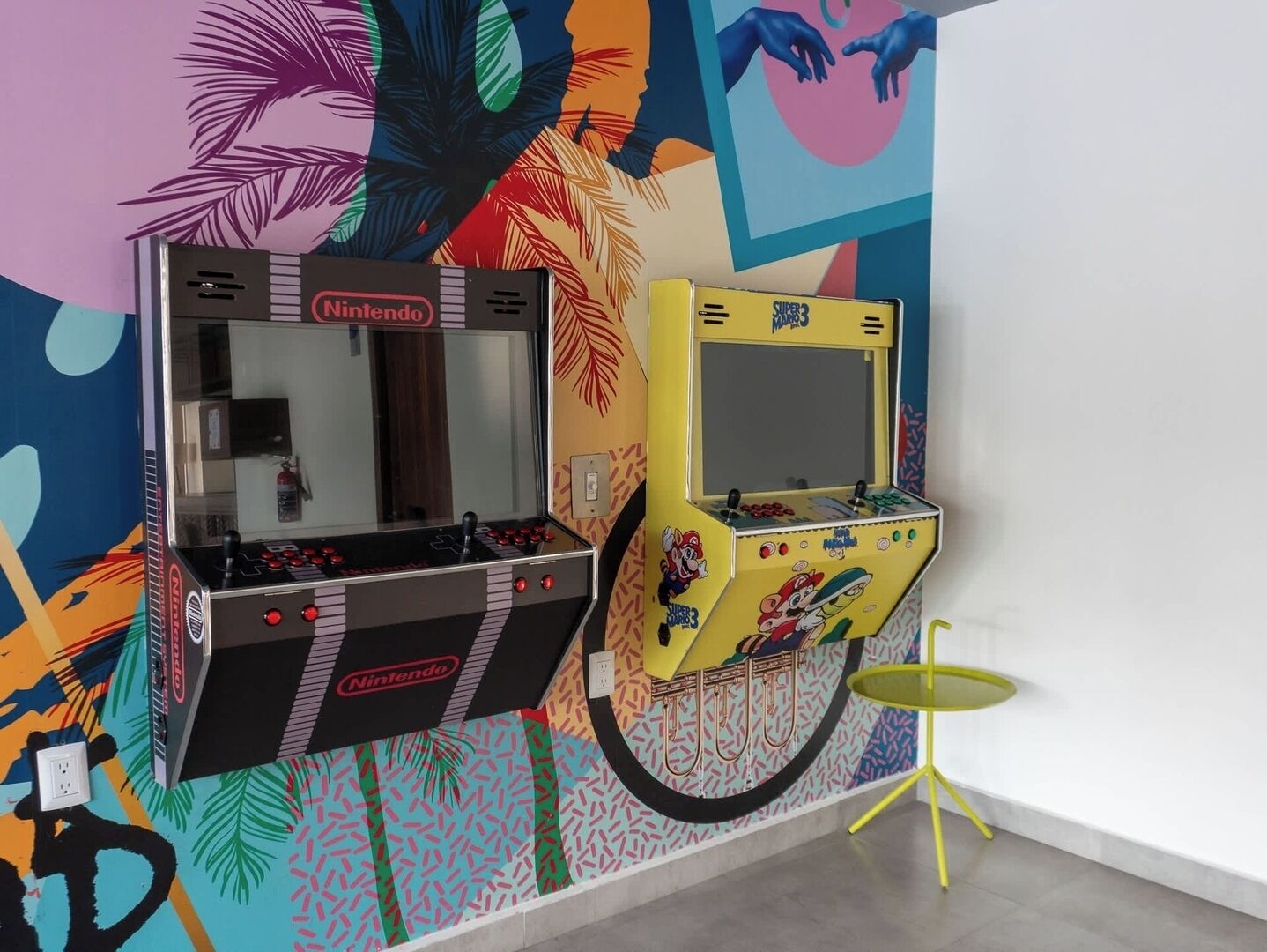 Twee speelautomaten, een zwart en een geel, met Nintendo-logo, voor een kleurrijke muur met abstracte patronen.