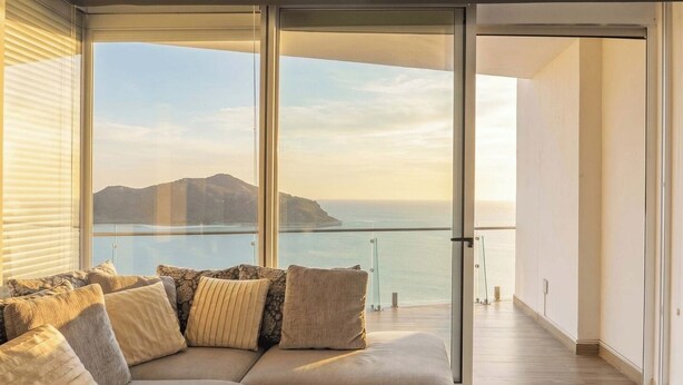 Ein modernes Wohnzimmer mit einer großen Couch, einer Glastür, die auf einen Balkon mit Blick auf das Meer und die Berge führt.