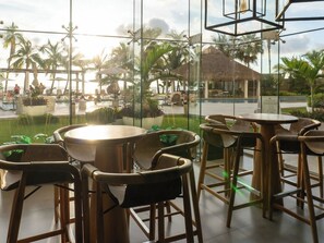 A glass-enclosed outdoor seating area with tables and chairs, overlooking a pool and palm trees. - Oceanfront Views of the Three Islands (Mazatlan)