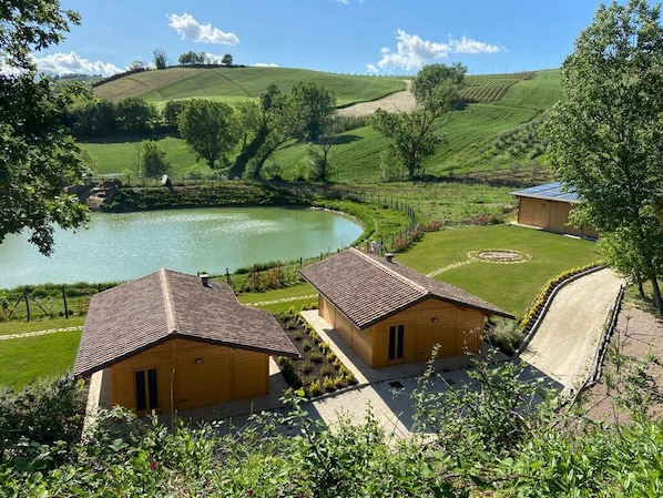 A rural setting with two wooden houses, a pond, and rolling hills in the background. - Wooden chalets for a stay in nature (Marche)
