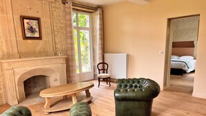 A room with a fireplace, a wooden bench, a green leather armchair, and a bed in the background. - Exceptional Manor House in Normandy – 6 En-suite Bedrooms – History & Elegance (Argences)