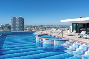 Une piscine sur le toit, accessible par des marches, entourée de chaises longues et offrant une vue sur la ville.