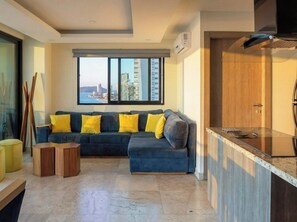 A modern living room with a sectional sofa, a small wooden side table, a kitchen area with a countertop, and a view of a cityscape through large windows. - Oceanfront Condo on the Malecón (Mazatlan)
