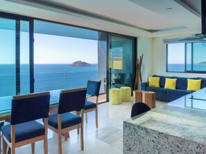 A modern living room with a glass wall overlooking the sea, a blue sofa with yellow cushions, and a dining area with chairs. - Oceanfront Condo on the Malecón (Mazatlan)